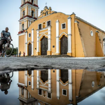 Église jaune avec son reflet dans une flaque d'eau et un passant à vélo.