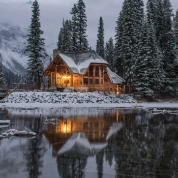 Chalet en bois éclairé au bord d'un lac entouré de sapins enneigés.