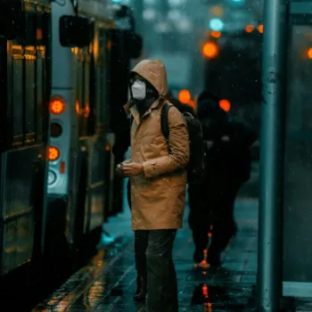 Une personne portant un manteau et un masque facial attend devant un bus sous la pluie.